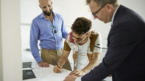 Salesman in suit watching as mid adult gay couple begins process of signing documents to finalize purchase in modern kitchen of their new home.